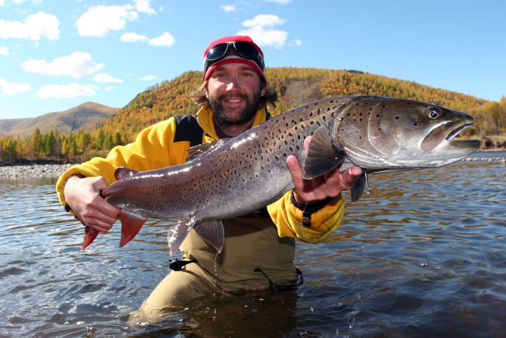 man with yellow coat and red hat holding a large fish while standing in a river