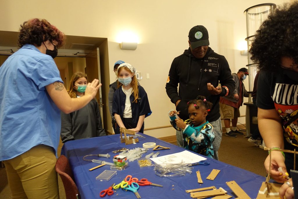 Families stand around a table with a blue table cloth, including a young girl, and use scissors, paper and glue.