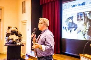 Dr. Thomas Adams wearing a button down and holding a mic. Behind him, the powerpoint reads "T.rex in Texas?" and a dinosaur skull sits to the left.