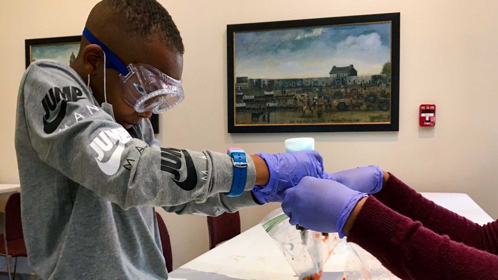 Child wearing goggles and gloves, focusing on pouring a liquid into a bag.