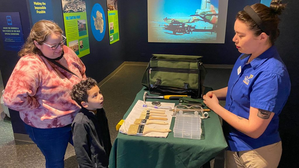 Mother standing behind son, who has an awed look on his face. They are looking at a table with paleontology tools.