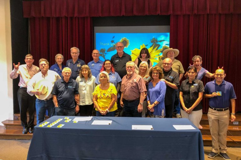 Approx. 20 adult volunteers stand behind a table and smile.