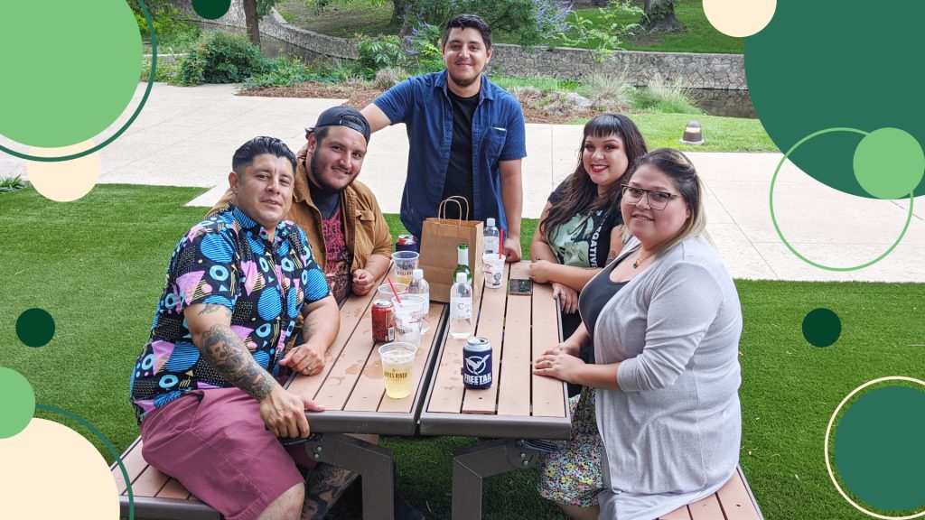 group of 5 smiles and sips beers while sitting at a picnic bench.